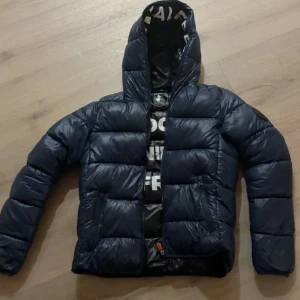 Mörkblå pufferjacka med huva - Säljer en mörkblå pufferjacka med huva och dragkedja framtill. Jackan har ett quiltat mönster och två sidofickor med dragkedja. Insidan av huvan har tryckt text i vitt för en cool detalj. Perfekt för kyliga dagar.