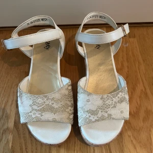 Vita sandaler med spets från Zoey 35 - Snygga vita sandaler från Zoey med bred rem i spets och glitterdetaljer. Sandalerna har en korkliknande plattformssula och justerbart spänne i guld.