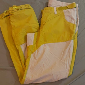 Gula och vita friluftsbyxor Haglöfs dam 38 - Snygga friluftsbyxor från Haglöfs i dammodell, storlek 38. Byxorna är gula med vita paneler och har praktiska detaljer som dragkedja och kardborre i midjan. Perfekta för utomhusaktiviteter under sommaren.