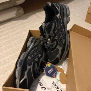 Asics - Säljer ett par snygga Asics sneakers i svart och grått med coola detaljer och meshpaneler för extra andningsförmåga. Skorna har en chunky sula och klassisk snörning. Perfekta för dig som gillar sportig stil och vill ha bekväma skor till vardagen.