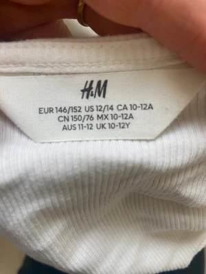 Vit ribbad linnetopp med spets H&M - Säljer en vit ribbad linnetopp från H&M i storlek 146/152. Toppen har smala axelband och v-ringning med söt spetskant upptill. Perfekt att styla med jeans eller kjol för en fräsch look.