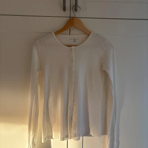 Vit ribbad kofta med knappar - Supersöt vit kofta från brandy  med ribbad struktur och diskret mönster. Rund halsringning, långa ärmar och knäppning framtill med små vita knappar. Perfekt att slänga över en topp när det är lite kyligt ute. Använt fåtal gånger