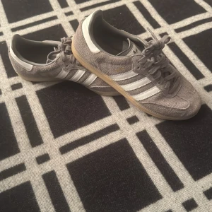Adidas Samba grå mocka sneakers - Adidas Samba sneakers i grå mocka med klassiska vita ränder på sidorna och gummisula. Skorna har snörning och rund tå, samt detaljer i vitt på hälen. Perfekt för dig som gillar retrostil och vill ha ett par snygga sneakers med skön passform.