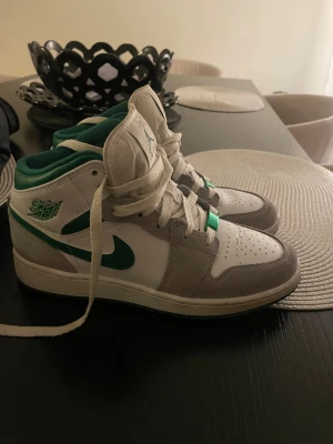 Nike Air Jordan 1 Mid vit/grön - Nike Air Jordan 1 Mid sneakers i vitt, grått och grönt med klassisk snörning och rund tå. Ovandelen är i skinn och mocka, med gröna detaljer på swoosh och logga. Skön platt sula och ikonisk Jordan-vibe. Perfekt för dig som gillar streetstyle.