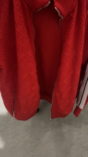 Röd kofta från Palm Angels - Palm Angels tröja i rött med vit och svart rand längs ärmarna. tröjan har ett snyggt geometriskt mönster i tyget, dragkedja framtill och ståkrage. Materialet känns som polyester och passformen är normal. Perfekt för dig som gillar streetwear.