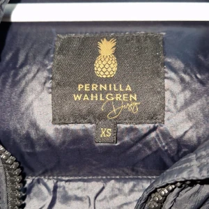 Svart pufferjacka Pernilla Wahlgren XS - Svart lång pufferjacka från Pernilla Wahlgren med huva och grova snören. Jackan har quiltad design, dragkedja framtill och två sidofickor. Materialet är glansigt och syntetiskt, perfekt för kalla dagar. Stilsäker look med guldiga detaljer på insidan.