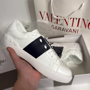 Valentino Garavani sneakers vit/marin blå - Säljer ett par snygga Valentino Garavani sneakers i vitt skinn med en bred marinblå läderdetalj över vristen och ikoniska nitar på hälen. Klassisk rund tå, platt sula och vita snören. Perfekta för dig som vill ha en clean men lyxig look. Kom med bud!