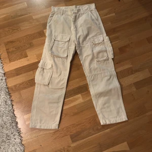 Beige cargopants från Ronning, storlek 28 - Säljer ett par beige cargopants från Ronning i storlek 28. Byxorna har flera stora fickor på sidorna och bak, klassisk cargostil och rak passform. Materialet är bomull och de stängs med dragkedja och knapp framtill. Perfekt för dig som gillar streetwear och funktionella detaljer.
