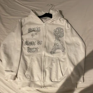 Vit hoodie med strasstenar från Blinglucid - Vit hoodie med dragkedja från Blinglucid, dekorerad med glittriga strasstenar och texten 'Paris is always the home' samt ett motiv av Eiffeltornet. Tröjan har huva och fickor framtill. Materialet är en mix av bomull och polyester.