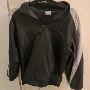 Svart och grå Nike vindjacka 158 xs - Nike vindjacka i svart med gråa paneler på ärmarna och röd swoosh på bröstet. Jackan har halv dragkedja, huva och meshfoder. Stor Nike-logga på ryggen. Perfekt för dig som gillar sportig och clean stil. Vintage