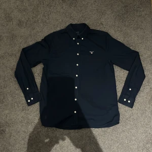 Mörkblå skjorta från GANT Barn - Klassisk mörkblå skjorta från GANT med vit broderad logga på bröstet och vita knappar. Skjortan har lång ärm, button down-krage och stilren look. Perfekt för dig som gillar en clean och tidlös stil.