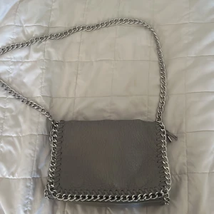 Grå axelväska med kedjedetaljer från Tiamo - Snygg grå axelväska från Tiamo i skinnimitation med grova silverfärgade kedjor runt kanterna och som axelrem. Väskan har dragkedja både på insidan och utsidan. OBS på sista bilden ser du lite rost vilket jag bara vill påpeka. (Inget som syns på långt håll)💗💗