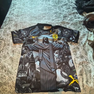 Svart Brasil Nike Dri-Fit fotbollströja - Unik svart Brasilien fotbollströja från Nike med Dri-Fit material. Framsidan har ett detaljerat konstnärligt tryck med Kristusstatyn, kyrkor och fåglar i grå och svart toner samt gula Brasil-loggor. Kortärmad och med rund hals. Perfekt för dig som vill sticka ut på planen.