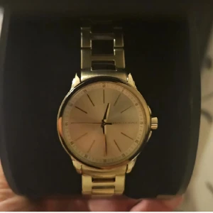 Guldig klocka från Armani Exchange använd 2 gånger  - Stilren klocka från Armani Exchange i guldigt metallarmband och rund urtavla. Klockan har minimalistisk design med guldfärgad urtavla och diskreta markeringar. Perfekt accessoar för dig som gillar klassisk och snygg stil.