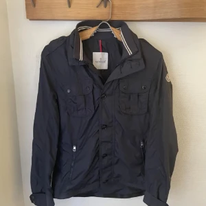 Marinblå vindjacka från Moncler - Snygg vindjacka från Moncler! Två stora bröstfickor med knappar och dragkedjor vid sidorna. Har haft den i ca 2 år utan defekter, STORLEK 3