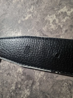 Svart skärp med silvrigt H-spänne - Snyggt svart bälte med silvrigt H-format metallspänne. Bältet har en slät yta med en lätt textur och insidan är ljusbeige. Perfekt för att ge din outfit en stilren och modern touch. Passar till jeans eller kostymbyxor och är lätt att matcha med olika looks.