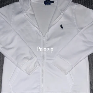Vit hoodie med zip från Polo Ralph Lauren - Vit hoodie från Polo Ralph Lauren med dragkedja och klassisk blå logga på bröstet. Tröjan har huva, långa ärmar och två fickor framtill. Perfekt för chill dagar och enkel att matcha med jeans eller joggers.