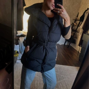 Svart pufferväst med bälte - Säljer en svart pufferväst med bred krage och midjebälte som ger en snygg siluett. Västen  är vadderad och har praktiska fickor på sidorna. Perfekt för dig som gillar streetstyle och vill hålla dig varm under kyliga dagar.