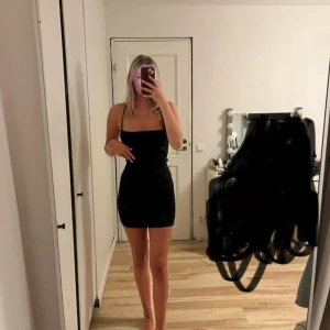 Svart kort klänning med snörning - Säljer en svart, tight och kort klänning med smala axelband och öppen rygg med snygg snörning. Klänningen har en stilren och enkel design som framhäver figuren. Aldrig använd 