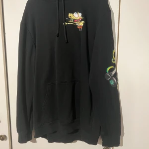 Svart Spongebob hoodie från H&M - Svart hoodie från H&M med Spongebob-motiv på bröstet och coolt tryck på ena ärmen med stekspade och färgglada detaljer. Tröjan har huva med snörning och stor magficka framtill. Regular fit och mjukt material som är skönt att ha på sig.