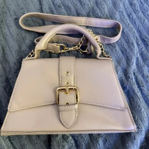 Beige axelväska med gulddetaljer - Snygg beige axelväska med både handtag och axelrem i kedja. Väskan har en dekorativ guldfärgad spänndetalj framtill och är tillverkad i slätt syntetmaterial. Perfekt storlek för det viktigaste och ger en stilren look.