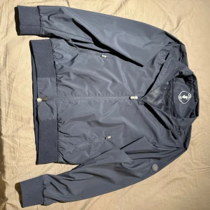 Blå vindjacka från Save The Duck - Snygg marinblå vindjacka från Save The Duck med ribbade muddar och krage. Jackan har två fickor med dragkedja, diskret logotyp på ärmen och meshfoder vid halsen. Bra skick,dessutom så är den mot vatten..