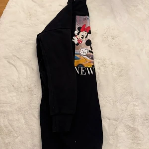Svart Disney sweatshirt från FB Sister - Cool svart sweatshirt med tryck av Minnie, Musse och Långben framför en New York-taxi. Tröjan har ribbade muddar och rund hals. Perfekt för dig som älskar Disney och streetstyle. Materialet är mjuk bomull och polyester.