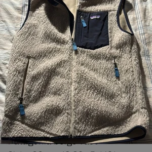 Beige fleeceväst från Patagonia M - Säljer en beige fleeceväst från Patagonia i storlek M. Västen har fluffig yta, marinblå detaljer och en bröstficka med dragkedja. Två sidofickor med blå zip-pullers och hel dragkedja framtill. Perfekt för lager-på-lager och chill stil.