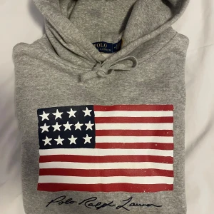 Grå hoodie Ralph Lauren - Snygg grå hoodie från Polo Ralph Lauren i storlek M. Framsidan har ett stort tryck med amerikanska flaggan i rött, vitt och blått samt Polo Ralph Lauren-loggan under. Tröjan har huva med snörning och är tillverkad i mjuk bomull. Perfekt för dig som gillar klassisk streetstyle.