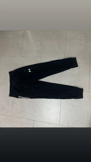 Svarta träningsbyxor Under Armour M - Svarta träningsbyxor från Under Armour i storlek M. Byxorna har elastisk midja med snörning, ribbade muddar vid benslut och vit UA-logga på benet. Tillverkade i lätt syntetmaterial som andas, perfekta för sport och chill.