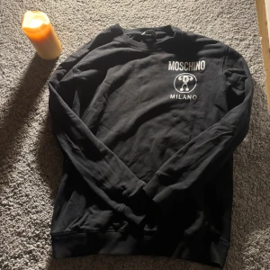 Svart Moschino sweatshirt med tryck - Svart sweatshirt från Moschino med vitt tryck på bröstet där det står 'Moschino Milano' och en ikonisk logga. Tröjan har rund halsringning, långa ärmar och ribbade muddar. Perfekt för dig som gillar stilrena och trendiga märkesplagg.