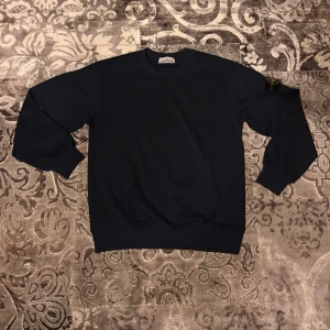 Svart Stone Island sweatshirt - Säljer en svart sweatshirt från Stone Island med klassisk rund hals och ribbade muddar. Tröjan har den ikoniska Stone Island-loggan på vänster ärm. Perfekt för dig som gillar stilrena och exklusiva plagg.