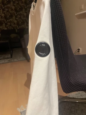 Vit sweatshirt från C.P. Company - Vit långärmad sweatshirt från C.P. Company med rund hals och klassisk passform. Tröjan har en ikonisk svart linsdetalj på vänster ärm och är tillverkad i mjuk bomull. Perfekt för dig som gillar stilrena och cleana plagg med en twist.