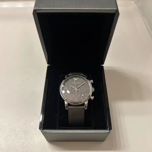Emporio Armani klocka - Snygg kronografklocka från Emporio Armani med rund urtavla i mörkgrått och silverdetaljer. Armbandet är i skinn och boetten är i blankt silverfärgat metall. Kommer i original ask med logga. Perfekt för dig som gillar stilrena accessoarer. Finns tecken på användning. 
