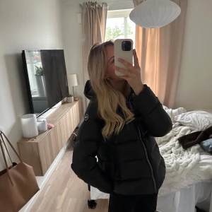 Svart Colmar pufferjacka med huva - Säljer en svart pufferjacka från Colmar med huva och snyggt, glansigt material. Jackan har en tydlig logga på ärmen, dragkedja framtill och två fickor med dragkedja. Perfekt för dig som vill ha en varm och stilren jacka med cool streetkänsla.