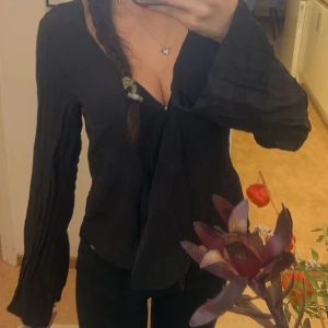 Svart omlottblus med v-ringning - Säljer en svart omlottblus med djup v-ringning och långa, vida ärmar. Blusen har en lätt skrynklig struktur och sitter snyggt med en loose passform. Perfekt för dig som gillar en chill och stilren look.