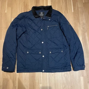 Mörkblå quiltad jacka från Tuxer - Quiltad mörkblå jacka från Tuxer med snygg krage i kontrastmaterial, praktiska fickor med tryckknappar och en bröstficka med dragkedja. Jackan har långärmad design och stilrena detaljer som ger en modern look. Perfekt för dig som gillar klassisk stil med edge.