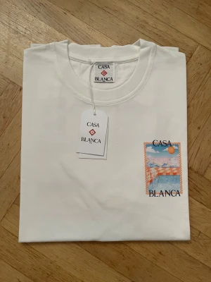Vit Casa Blanca t-shirt med tryck - Snygg vit t-shirt från Casa Blanca i 100% ekologisk bomull. Framtill finns ett färgstarkt tryck med sol, moln och hav i orange och blå toner samt logotypen. Klassisk rund hals och korta ärmar. Perfekt för dig som gillar stilrena plagg med unik detalj.