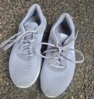 Grå sneakers från Nike - Snygga grå Nike sneakers med vita detaljer. Skorna har snörning och en platt sula med tydligt Nike-logo under foten. Perfekt för dig som gillar sportig stil och vill ha lätta skor i syntetmaterial.