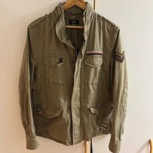 Beige fieldjacket från Mason's - Snygg beige overshirt från Mason's med coola detaljer som broderade märken på bröstfickan och ärmen. Jackan har flera fickor med lock och knappar, samt en upphöjd krage. Perfekt för dig som gillar street och utility vibes. Nypris runt 5000 kr