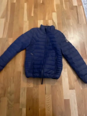 Mörkblå dunjacka från Parajumpers - Säljer en snygg mörkblå dunjacka från Parajumpers med klassisk patch på ärmen. Jackan har quiltad design, dragkedja framtill och stårkrage. Insidan är vit och materialet är lätt och skönt, perfekt för kyliga dagar. Passar dig som vill ha en stilren och funktionell jacka.