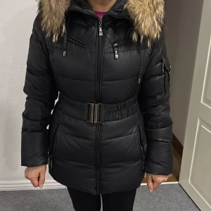 Svart pufferjacka med päls från Hollies - Svart pufferjacka från Hollies med snygg päls på huvan, bälte i midjan och flera dragkedjor. Jackan har lång ärm, ficka på armen och mönstrat innerfoder. Perfekt för kalla dagar med sin tjocka och fluffiga look. Jackan är använd 2 vintrar men ser helt ny och fräsch ut. Den är i storlek 38. 