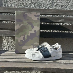 Valentino Garavani Open sneakers vit/blå - Säljer ett par Valentino Garavani Open sneakers i vitt skinn med bred mörkblå skinnpanel över snörningen. Klassisk rund tå och platt sula, med coola nitar på hälen. Kommer med box i camouflage-mönster. Perfekt för dig som gillar lyxiga streetwear vibes.
