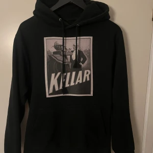 Svart hoodie Kellar The Cool Elephant - Svart hoodie med stor Kellar-print på framsidan och liten The Cool Elephant-logga på ryggen. Tröjan har huva med snörning och en klassisk känguruficka framtill. Perfekt för dig som gillar unika tryck och streetwear.