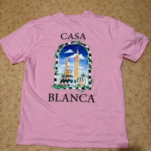 Casa Blanca rosa t-shirt med tryck - Snygg rosa t-shirt från Casa Blanca med stort färgglatt tryck på ryggen som föreställer ett fönster med palmer och torn. T-shirten har klassisk passform och rund hals. Perfekt statement-plagg för dig som gillar unika prints.