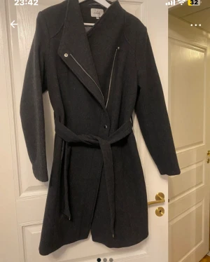 Mörkgrå kappa från Isolde med bälte - Snygg mörkgrå kappa från Isolde i storlek 38. Jackan har dragkedja och matchande bälte i midjan. Långärmad och tillverkad i ullblandning, perfekt för kyliga dagar. Stilren och enkel att matcha med olika outfits.