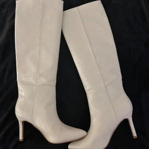 Kräm vita höga stövlar med klack - Säljer ett par snygga krämvita stövlar med spetsig tå och hög klack. Stövlarna har ett stilrent croco-mönster och är tillverkade i skinnimitation. De går upp till knäet och har en elegant, smal siluett som ger en trendig look.