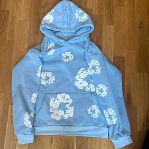  denim tears hoodie - Säljer en ljusblå hoodie med huva och stora vita molnliknande mönster över hela tröjan. Tröjan har en klassisk känguruficka framtill och ribbade muddar vid ärmslut och nederkant. Perfekt för dig som gillar chill och unik streetstyle.