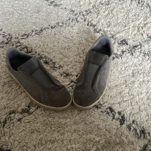 Grå slip-on sneakers från Axel Arigato - Snygga grå slip-on sneakers från Axel Arigato med stilren design och diskret logga i guld på sidan. Skorna har en rund tå, platt sula och är tillverkade i mjuk mocka med elastiskt band över vristen för enkel påtagning. Perfekta för dig som gillar minimalistisk stil.