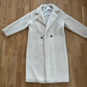 Vit teddyjacka med krage - Säljer en lång, vit teddyjacka med bred krage och två svarta knappar framtill. Jackan har en rak och oversized passform med mjukt, fluffigt material som ger en mysig vibe. Behövs tvättas då de finns lite fläckar, men försvinner lätt med en tvätt